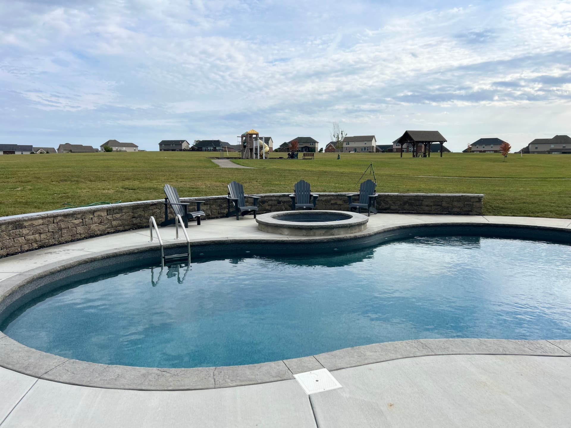 Curved pool with concrete deck, seat wall and round fire pit with Adirondack chairs